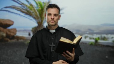 Elinde kitap olan papaz cüppeli bir adam sahil kenarında palmiye ağaçları ve okyanus manzaralı bir yerde durup parlak bir anı işaret ediyor.