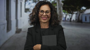 Sokakta gülümseyen bir kadın, dışarıda tablet kullanırken, siyah giyinmiş, gözlüklü, arka planda bulanık bir şehir ortamında olgun bir güven sergiliyor..