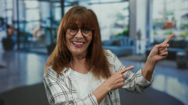 Üst düzey İspanyol kadın gülümseyerek ve kapalı bir otel lobisinde gözlük ve ekose gömlek takarak neşeli ve davetkar bir atmosfer sunuyor..