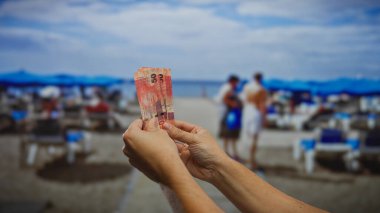 Arka planda plaj sandalyeleri ve deniz olan Güney Afrika Rand 'i tutan adam güneşli bir günde tatil harcamaları öneriyor..