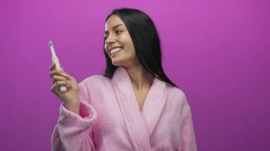 Pembe bornozlu genç İspanyol kadın elektrikli ve geleneksel bir diş fırçasını canlı pembe bir duvar arkasına tutarken gülümsüyor..