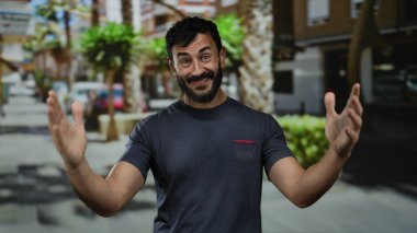 Sakallı İspanyol bir adam şehir caddesinde ağaçlar ve binalar arkasında mutluluk ve kendine güveni ifade ederek neşeli bir şekilde dolaşır..