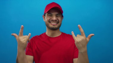 Stüdyoda parlak kırmızı şapkalı ve tişörtlü genç adam rock boynuzlarını diliyle gösteriyor..