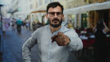 Canlı sokak sahnesinde sakallı İspanyol bir adam masaların ve yayaların hareketli kentsel geçmişleri arasında gözlük ve gri gömlek takıyor..