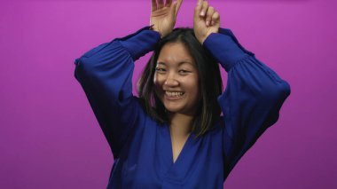 Genç Çinli kadın neşeli bir şekilde gülümsüyor canlı pembe bir arka plana karşı boynuz hareketi yapıyor, neşe ve eğlenceyi ifade ediyor..