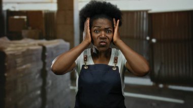 İnşaat alanında önlük giyen bir kadın stresli görünüyor. Kafasını kapalı bir endüstriyel ortamda tutuyor. Bu da işle ilgili bir hayal kırıklığı ya da kaygı olduğunu gösteriyor..