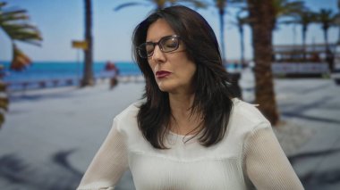 Orta yaşlı İspanyol kadın sandığı deniz kenarında güneşli gökyüzünün altında palmiye ağaçlarıyla kaplı bir sandık; kalp krizi acısı.