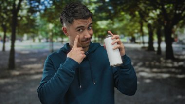 Parkta elinde grafiti ifadesi olan beyaz sprey bir kutu tutan, mavi kapüşonlu, ağaçların ve çimenlerin bulanık arka planına yaslanmış genç bir İspanyol.