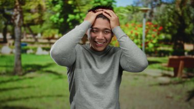 Parktaki genç Çinli adam, verimli yeşillikler ve güneşli bir günde park banklarıyla çevrili çeşitli duygular gösteriyor..