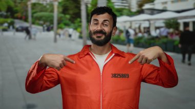 Hapishane tulumu giyen Hispanik bir adam işlek bir caddede, kentin dış manzarasıyla çevrili bir sırıtışla kendine bakıyor..