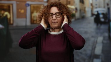 Kıvırcık saçlı kadın şehir sokaklarında kulaklarını elleriyle kapatıyor, gözlük ve kestane rengi kazak giyiyor, hayal kırıklığını ya da gürültüyü gösteriyor..