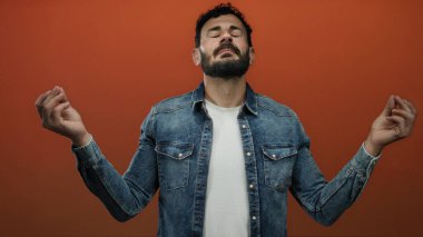 İspanyol bir adam turuncu bir arka plan karşısında sakal ve kot ceketle meditasyon yapıyor. Dua eden eller, gözleri kapalı, sükunet ve farkındalık sergiliyor.