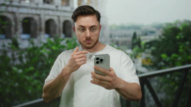 Sakallı genç adam Roma kolezyumunun yanında dikilirken akıllı telefona şaşırdı, arka planda yeşillik olan açık hava sahnesini gün ışığında çekti..