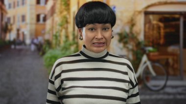 Çince kadın, bulanık arka planlı, çizgili süveter ve küpe giyen, kentsel bir sokak ortamında rahatsızlık gösteriyor, yüz ifadesi huzursuzluğu ve tereddüdü yansıtıyor..