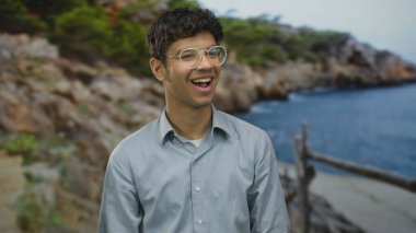 Gözlüklü bir adam açık mavi gökyüzü altında güneşli sahil şeridinde geniş bir gülümseme; tasasız mutluluk.