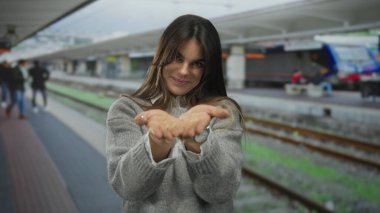 Gri kazaklı genç İspanyol kadın tren istasyonunda gülümsüyor. Tren rayları ve platformları görünüyor. Açık hava tren istasyonunda sıcak bir karşılama sahnesi yaratıyor..