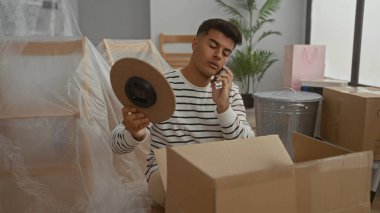 Çizgili gömlekli genç adam, yeni bir apartman dairesinde karton kutuları açarken telefonla konuşuyor. Yeni bir taşınma ya da taşınma işlemi öneriyor..