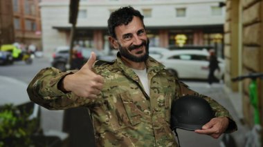 İspanyol üniformalı bir adam kalabalık bir sokakta elinde miğfer tutarak gülümsüyor. Bu da kendine güveni ve açık havada pozitif tutumunu gösteriyor..