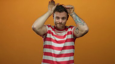 Çizgili tişörtlü genç İspanyol, izole edilmiş turuncu arka plana karşı neşeli kulak hareketi yapıyor..