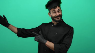 Sakallı İspanyol erkek aşçı canlı yeşil bir fon karşısında şakacı bir şekilde yan yatmış, siyah şef kıyafeti ve uyumlu bir şapka giymiş, aşçılık hevesini somutlaştırıyor..