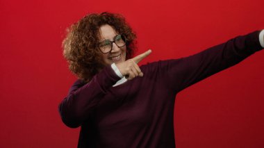 Kıvırcık saçlı ve gözlüklü, kendine güveni tam bir jest sergileyen canlı kırmızı arka planda gülümseyen bir kadın..