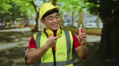 Açık hava parkında güvenlik önlüğü takan ve kasketli genç bir adam güvenle gülümseyen elektronik bir sigara tutuyor..