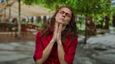 Kırmızı gözlüklü ve gömlekli bir kadın yeşil ağaçlar ve bulanık binalarla çevrili güneşli bir parkta dua ediyor..