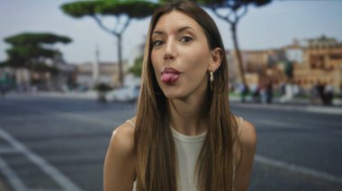 Uzun kahverengi saçlı, beyaz kolsuz bluzlu genç kadın şehir sokaklarında dil uzatıyor, arsız oyuncu tavırlar sergiliyor..