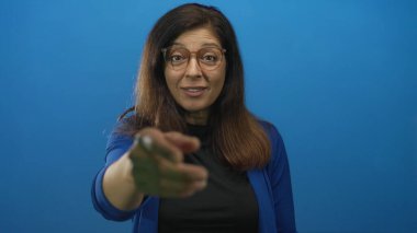 Gözlüklü ve uzun saçlı bir kadın mavi bir stüdyoda kamerayı işaret ediyor..