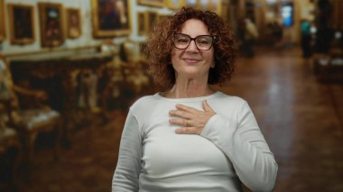 Orta yaşlı esmer bir kadın sanat galerisinde gülümsüyor, gözlük takıyor ve beyaz bir gömlek giyiyor..