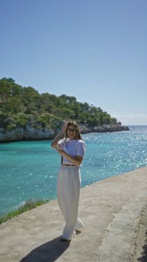 Santanyi 'deki güneşli plajda okyanus manzarasını yakalayan bir kadın, açık mavi gökyüzü altında canlı turkuaz su ve yemyeşil yemyeşil bir Mallorca..