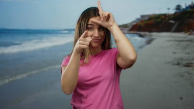 Ezik işareti yapan ve güneşli bir sahil sahilinde pembe gömleğiyle kamerayı işaret eden genç bir kadın.