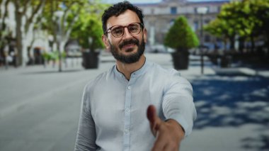 Gözlüklü İspanyol adam, güneşli bir sokakta, arka planda modern bir şehir ortamında yeşil ağaçların oluşturduğu dostça bir selamlaşmayla elini uzatıyor..
