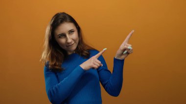 İzole edilmiş turuncu duvar arkaplanını işaret eden genç bir kadın mavi süveter giyiyor merak ve nişan ifadesi içeren bir yön ya da fikir kavramını gösteriyor