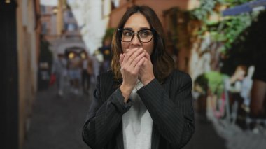 Kalabalık bir şehir caddesinde gözlüklü İspanyol kadın, bulanık insanlar ve kentsel çevre ile çevrili ağzı şaşkınlıkla kaplayarak, canlı bir şehir manzarası yaratıyor..