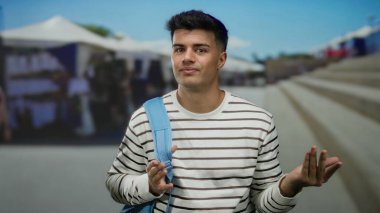 Şehir caddesinde mavi sırt çantalı genç bir adam açık havada çizgili gömlek giyiyor güneşli bir günde düşünceli görünüyor satıcıların ve basamakların bulanık geçmişiyle.