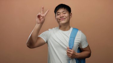 Mavi sırt çantalı genç Çinli adam gülümsüyor ve izole edilmiş kahverengi arka plan duvarına barış işareti yapıyor..