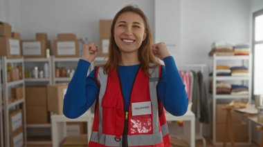 İç mekan hayır kurumunda gönüllü olarak gülümseyen genç bir kadın. Kırmızı yelek giyiyor. Etrafı düzenli kutular, malzeme ve kıyafetlerle dolu..