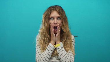 Sarışın bir gencin stüdyo resminde mavi arka planda sersemlemiş bir ifadeyle ellerini kullanan bir kadın..