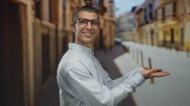 Beyaz adam kollarını açmış gülümsüyor gözlük ve çizgili gömlek giyiyor açık bir sokak arkaplanında.