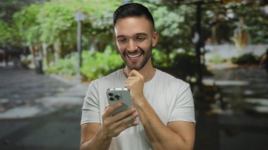 Yeşil bir parkta akıllı telefona gülümseyen, güneşli bir günde açık havanın tadını çıkaran, doğanın kucağında bir mutluluk ve rahatlama anı yakalayan genç bir adam..