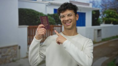 Beyaz binanın önündeki gülümseyen adam pasaport ve pasaportu tutuyor; seyahat heyecanı.