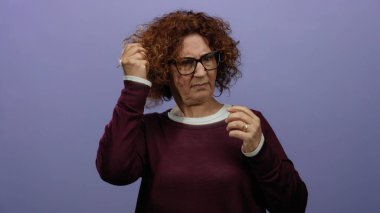 Kıvırcık esmer saçlı gözlüklü bir kadın mor izole edilmiş bir arka plana işaret ediyor. Kafa karışıklığı ya da tefekkür gösteriyor..