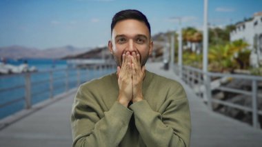 Yüzünde elleri olan genç bir adam güneşli bir günde arka planda deniz ve kumsal gezintisinde sürpriz yapıyor..