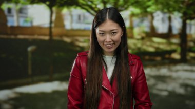 Parkta gülümseyen kırmızı ceketli bir kadın, güneşli bir günde, bulanık yeşil bir arka planda uzun saçlarını gösteriyor..