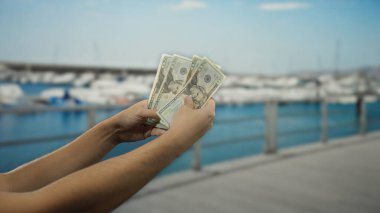 Sahilde bir liman kenarında Amerikan doları tutan ve arka planda görünen tekneleri olan bir adam, denizcilik ortamında finansal seyahat temalarını gözler önüne seriyor..