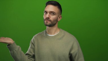 Latin kökenli sakallı genç adam elini uzatıyor ve bej bir süveter giymiş, koyu yeşil bir arka plana karşı derin düşüncelere dalmış bir ifade sergiliyor..