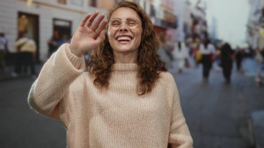 Kadın gülüyor ve el sallıyor kalabalık bir şehir caddesinde, arka planda bulanık insanlarla neşe ve şehir yaşamını yakalıyor ve gözlük takıyor ve rahat bir süveter giyiyor..