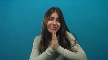 Elleri göğsünün önünde sıkıca bağlı olarak dua eden bir kadın saf mavi bir arkaplan karşısında, umutlarını veya umutlarını ifade eden, sakin ve basit bir düzende dikkat dağıtıcı şeylerden soyutlanmış bir kadın..