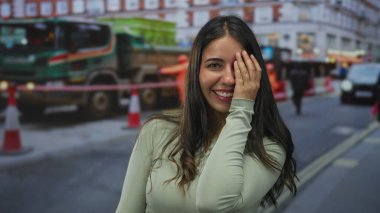 Bir şehir caddesinde, arkasında trafik konileri olan inşaat araçlarının önünde eli ile gözlerini kapayan genç bir kadın; çekingenlik.